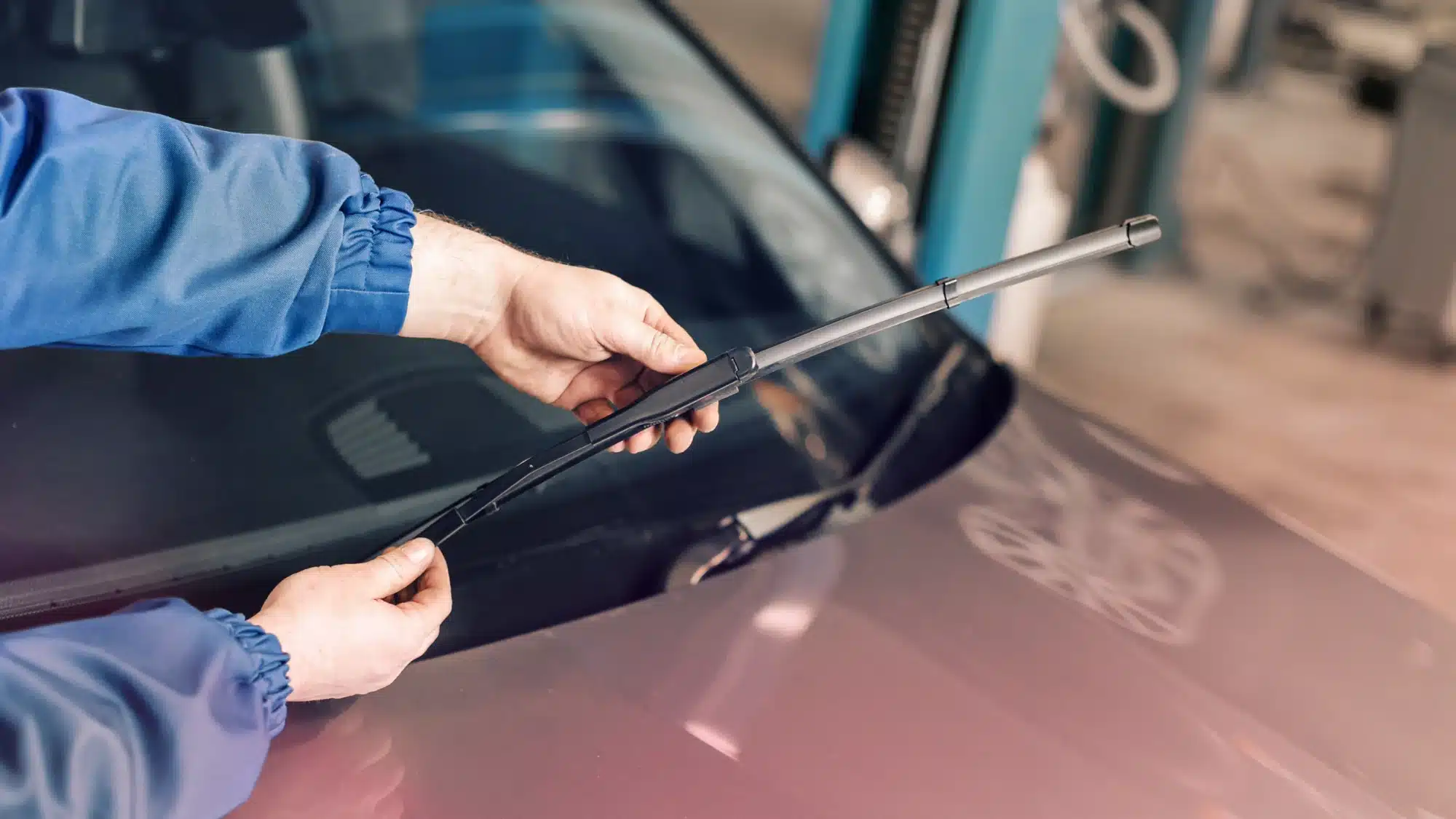 A mechanic in uniform inspecting a car's windshield wipers in a repair shop, with tools visible.